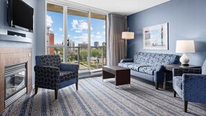A hotel room with a large window, a flat-screen TV, a sofa, two patterned armchairs, a coffee table, and a fireplace. - Victoria | Group Harbor Retreat | Pool + Harbor | 2BR (Victoria)