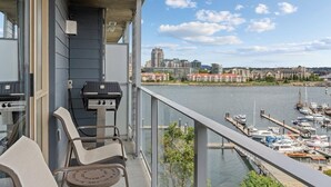 A balcony with a grill, chairs, and a view of a marina and cityscape. - Victoria | Group Harbor Retreat | Pool + Harbor | 2BR (Victoria)