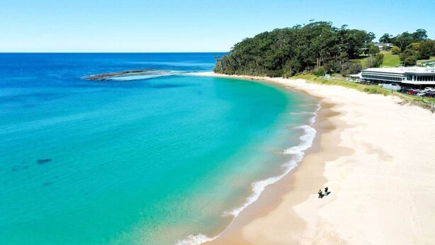 Een strand met helder turquoise water, een zandstrand en een gebouw met een plat dak aan de rechterkant.