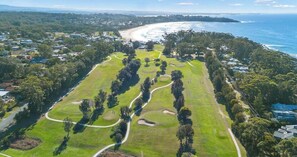 Luftaufnahme eines Golfplatzes mit Küstenlandschaft im Hintergrund, umgeben von Wohngebieten.