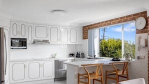 A modern kitchen with white cabinetry, a built-in microwave, and a large window with a view of greenery. - Bella Vista 1 - 3 bedroom Apartment Ocean View (Mollymook)