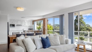 A modern living room with a large sofa, a dining area, and a kitchen with white cabinets and a brick accent wall. - Bella Vista 1 - 3 bedroom Apartment Ocean View (Mollymook)