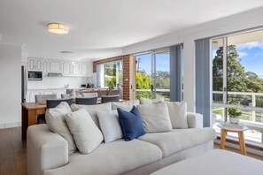 Ein modernes Wohnzimmer mit einem großen Sofa, einem Essbereich und einer Küche mit weißen Schränken und einer Wand in Backsteinoptik.