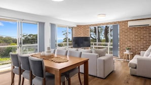 A modern living room with a dining area, large windows, and a brick wall. - Bella Vista 1 - 2 Bedroom Apartment Ocean Views (Mollymook)