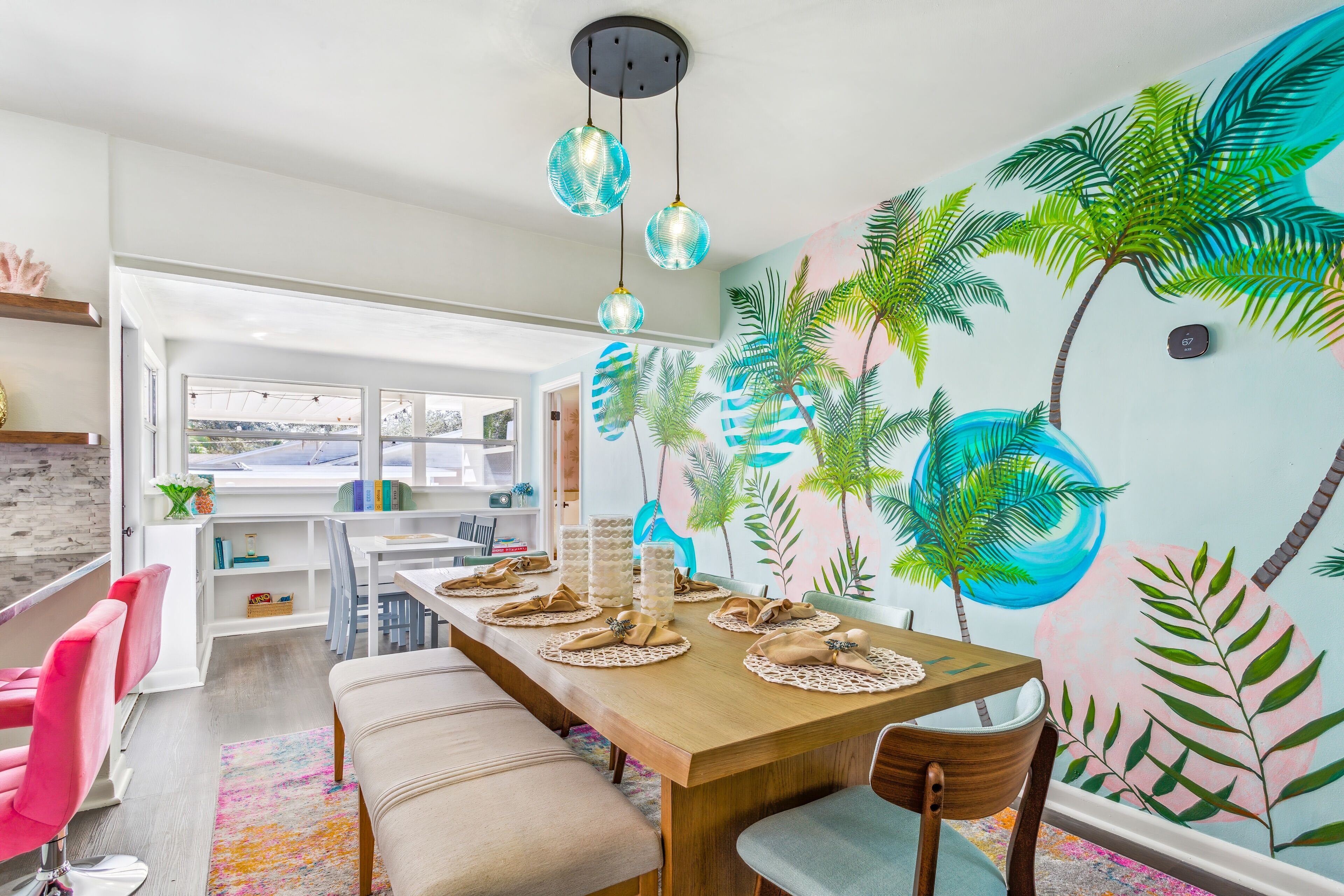 Un coin repas avec une table en bois dressée pour quatre personnes, un banc et une fresque représentant des palmiers sur le mur.