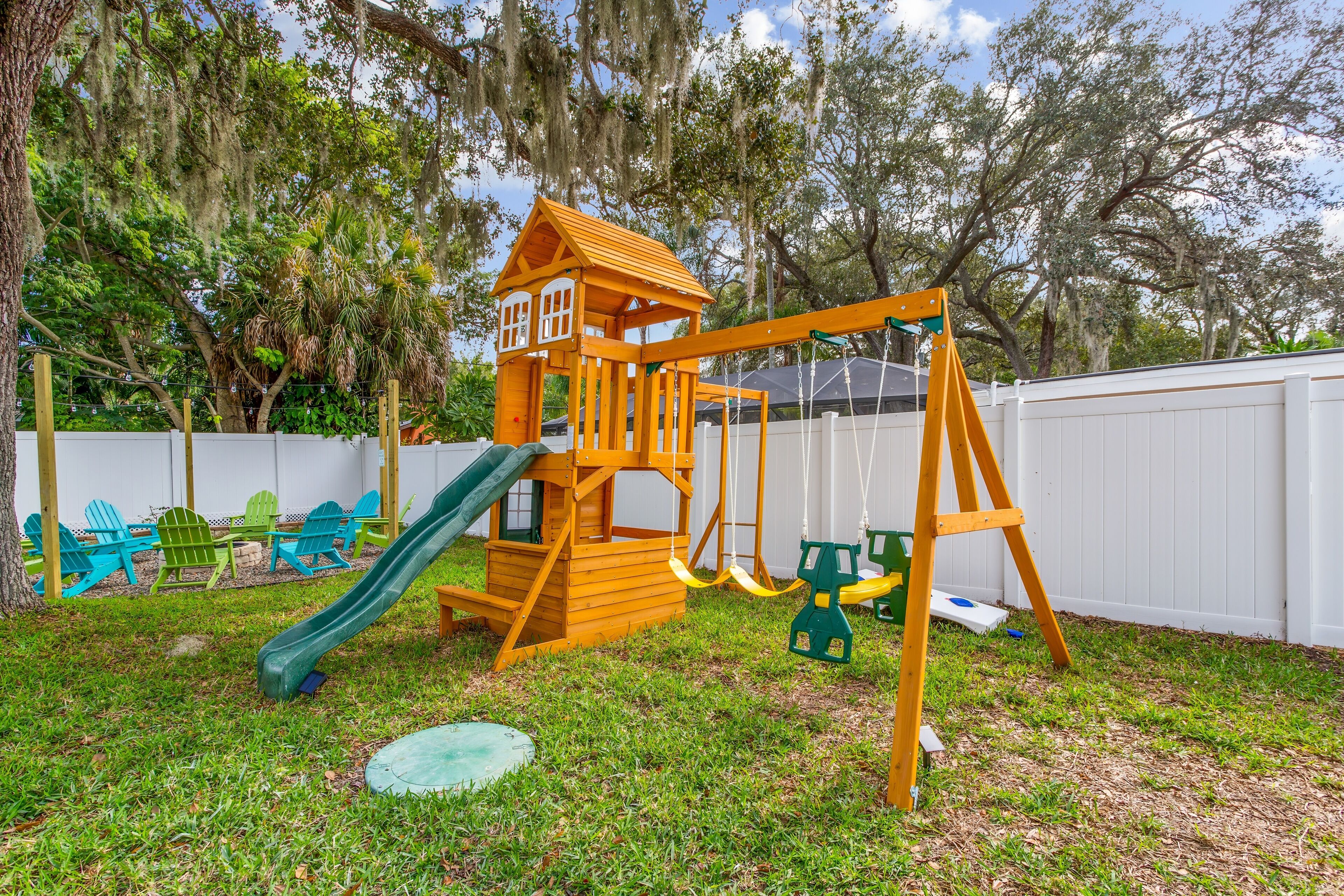 A wooden play structure with a slide and swings, surrounded by a white fence and trees.
