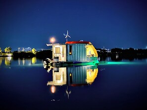 Ein festliegendes Boot mit einer kleinen Kabine, einer Windturbine und einer ruhigen Wasseroberfläche, die die Struktur spiegelt.