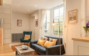 A living room with a sofa, armchair, and a table with a bowl. There are windows with curtains, a radiator, and framed pictures on the walls. - Roomy 2 Bed Garden Flat Quick Links to Central (London)