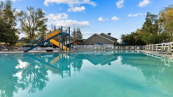 A clear swimming pool with a blue and yellow slide, surrounded by trees and a building with a white fence.