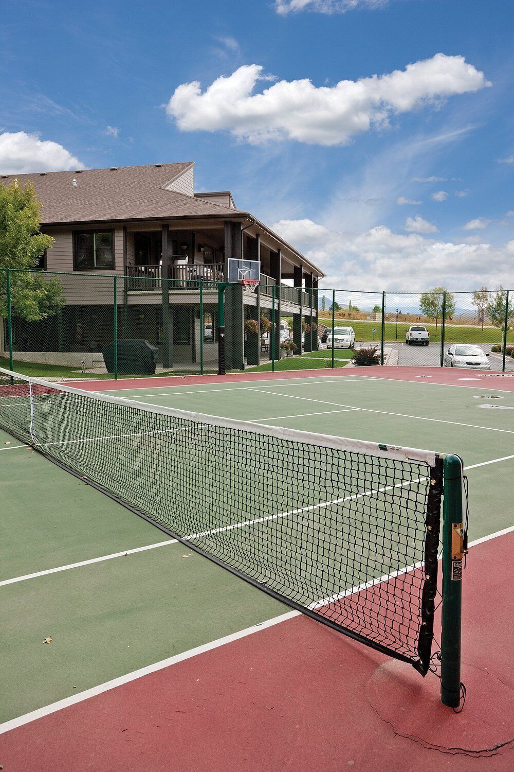 A tennis court with a net, green and red playing surfaces, and a building with a balcony in the background.