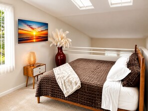 Ein Schlafzimmer mit einem Bett, einem Nachttisch, einer Vase mit weißen Blumen, einem Sonnenuntergangsbild und einem Fenster mit Jalousien.