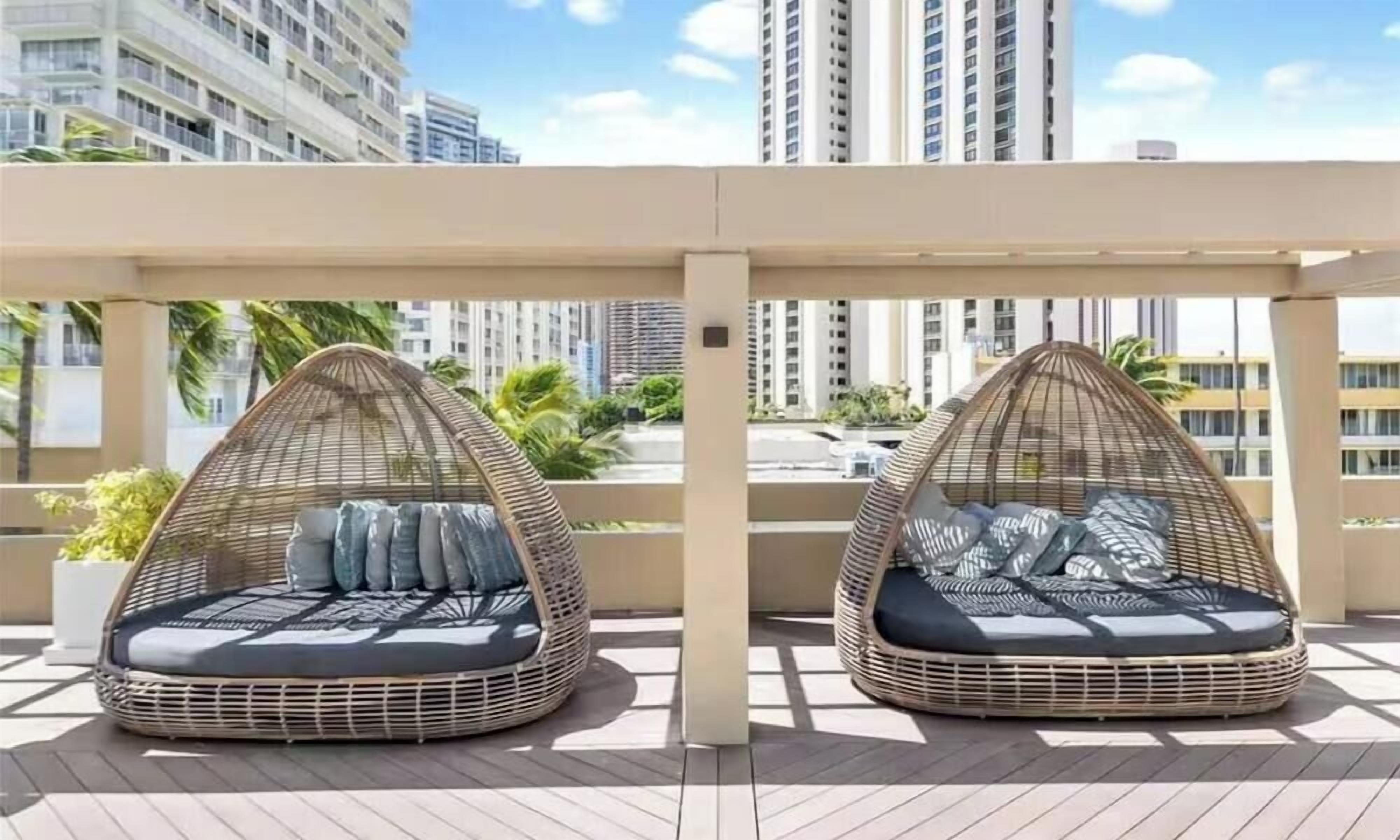 Two wicker hanging chairs with cushions on a balcony with a view of tall buildings.