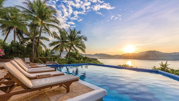 Una piscina infinita con sillas de descanso, palmeras y vistas al atardecer.