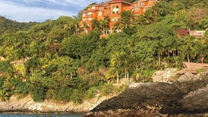 A coastal hotel with multiple floors, surrounded by lush greenery and a rocky shoreline. - Zihuatanejo | Compact Ocean Retreat | Pool + Beach (Zihuatanejo)