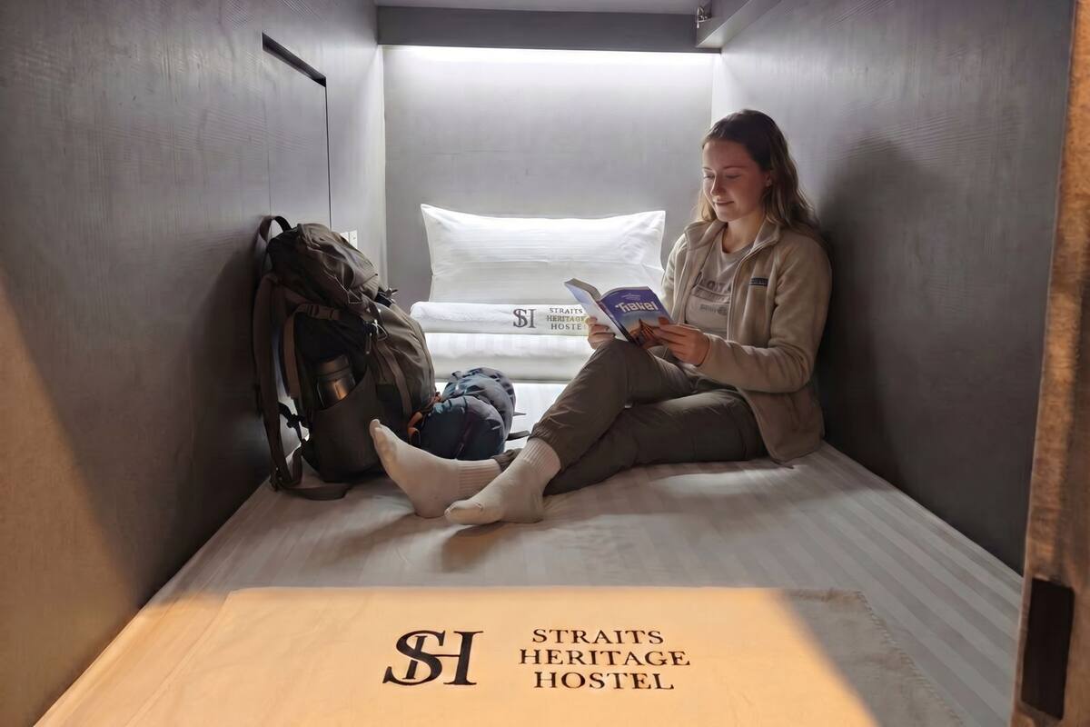 Une personne est assise sur un lit dans une petite pièce, avec un sac à dos et un livre, et un panneau au sol indique « STRAT’S HERITAGE HOSTEL ».