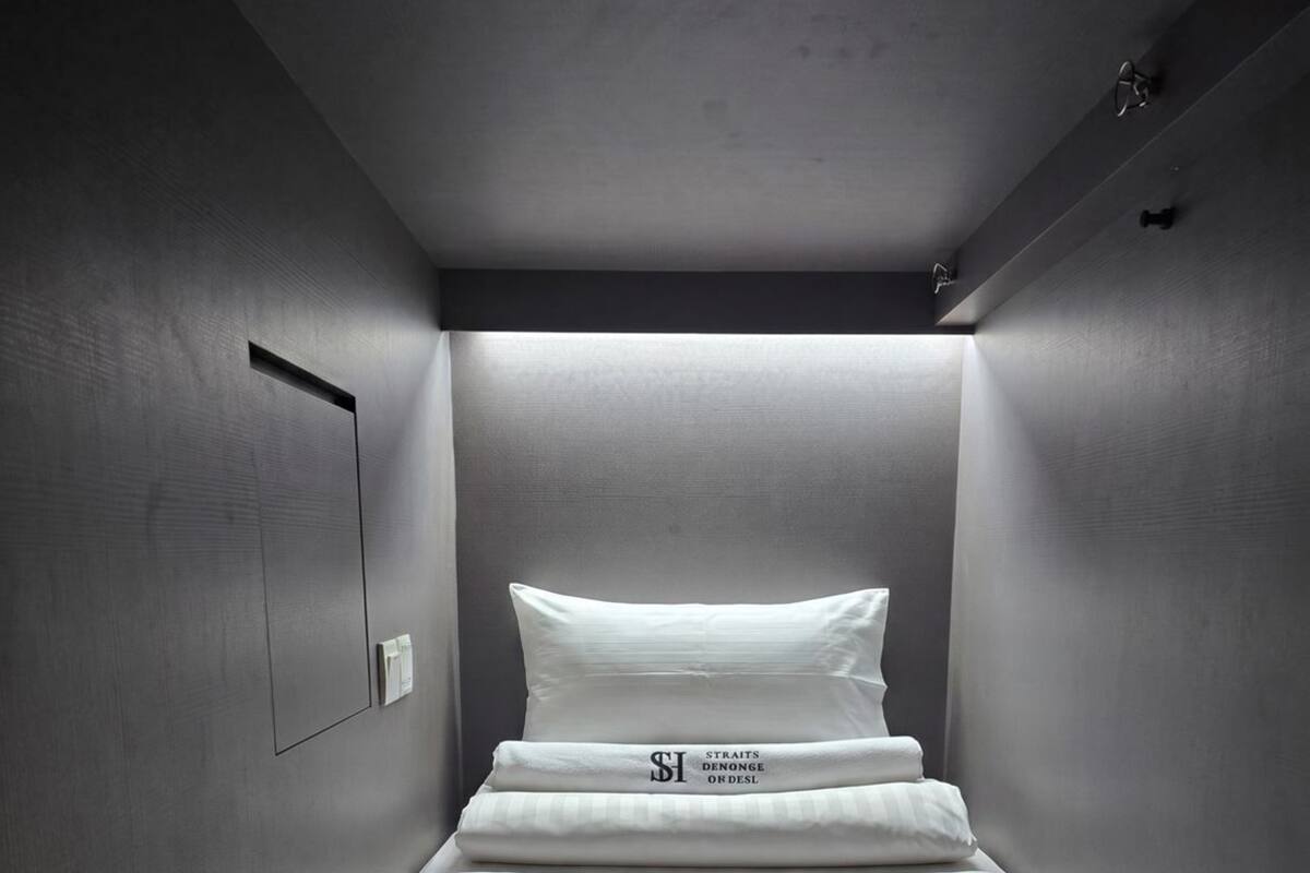 Une petite pièce étroite, avec un lit simple, une lampe fixée au mur et une porte.