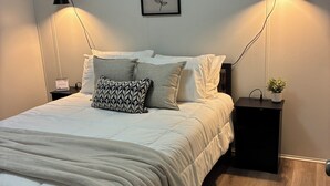 Un dormitorio ordenado con cama, dos mesitas de noche, una lámpara de pared, una pequeña planta en maceta y una obra de arte enmarcada.