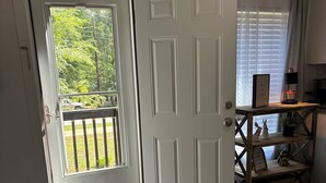 Una habitación con piso de madera, una puerta blanca, un balcón, una mesita de noche con una lámpara y un sofá.