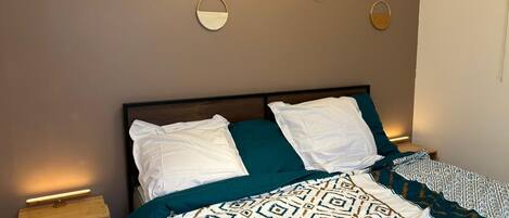Ein ordentlich bezogenes Bett mit weißer und türkisfarbener Bettwäsche, einer gemusterten Tagesdecke und einem Nachttisch aus Holz mit einer Lampe.