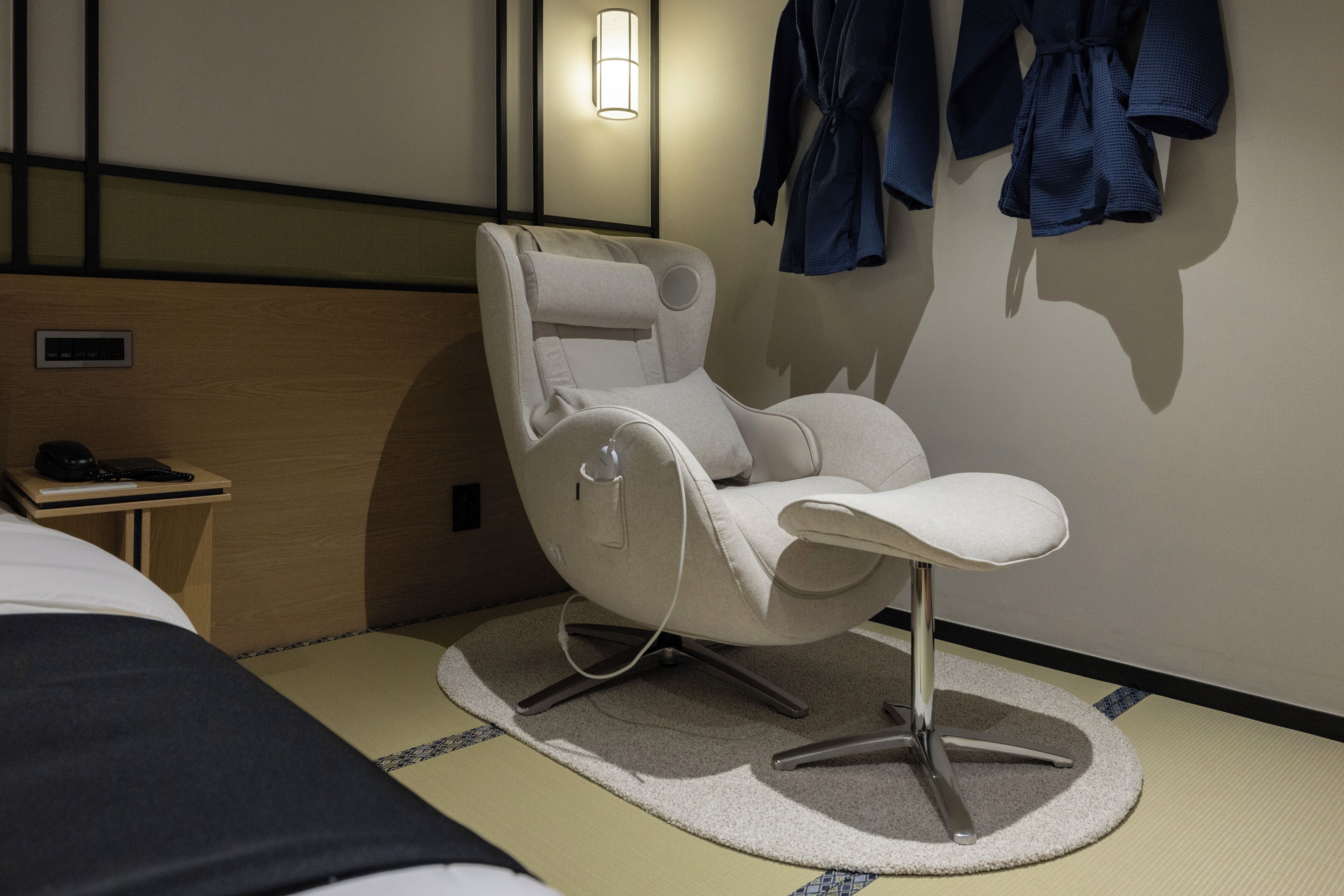 A modern hotel room with a massage chair, a bedside table with a phone, and a wall-mounted robe rack.