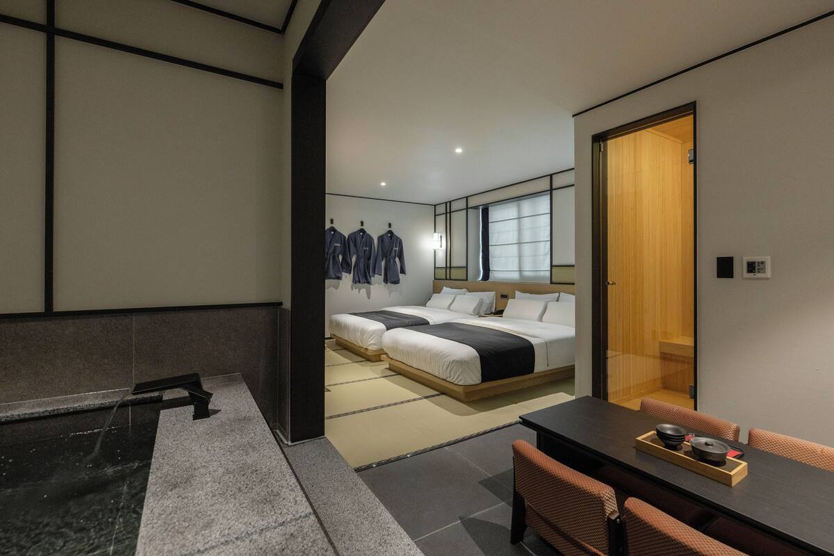 Quarto de hotel moderno com uma cama, uma mesinha com bandeja, um espaço de estar e vista para outro quarto com janela.