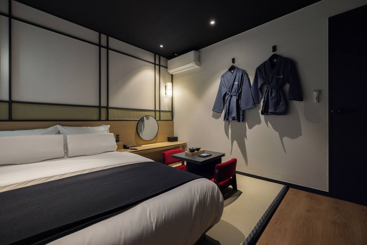 Quarto de hotel com uma cama, uma escrivaninha, uma cadeira e dois roupões pendurados na parede.