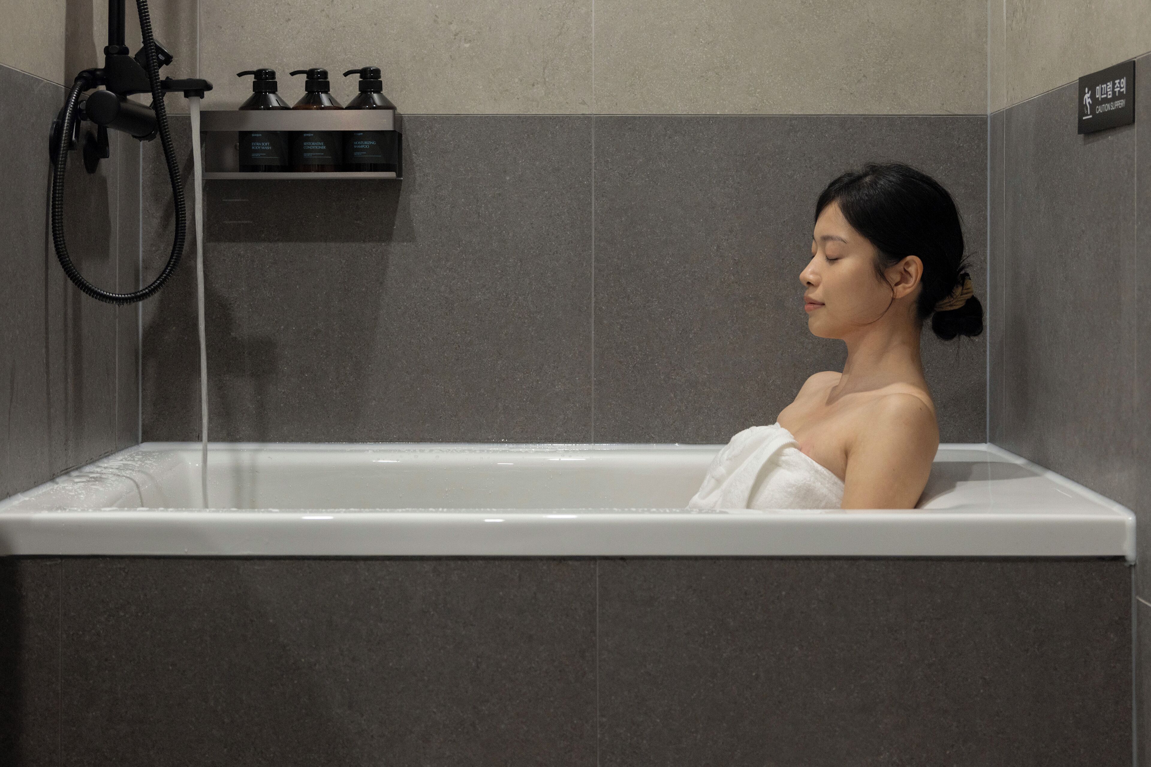 A person in a bathtub with a showerhead and toiletries on a shelf.