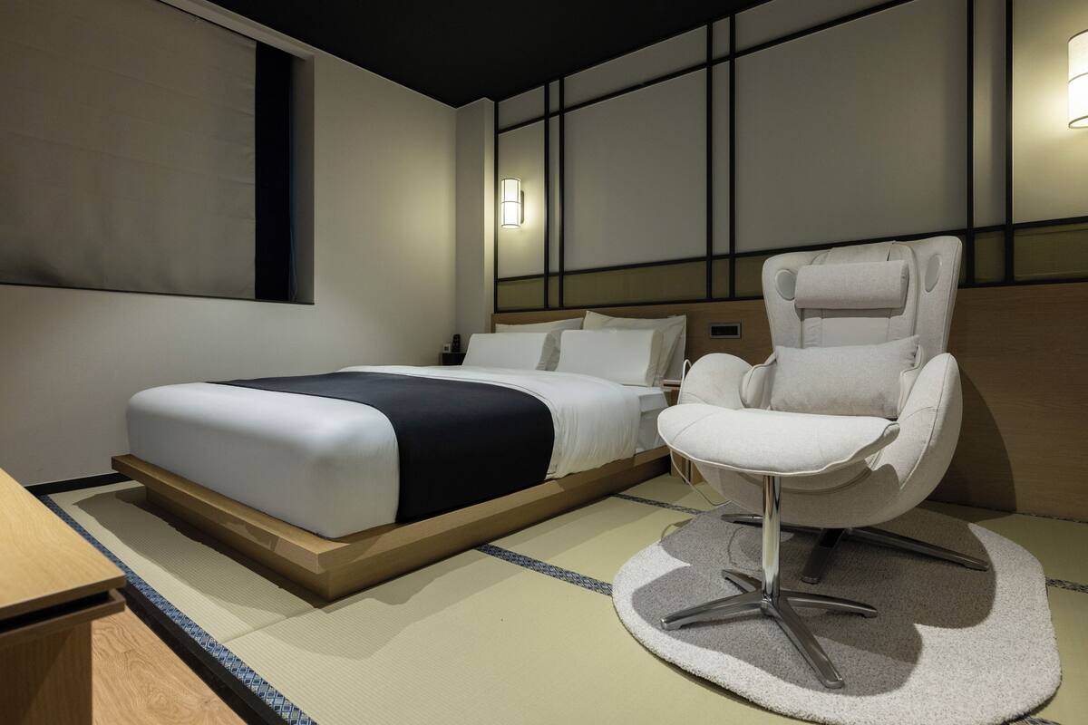 Quarto de hotel moderno com cama, uma cadeira e luminária fixada na parede.