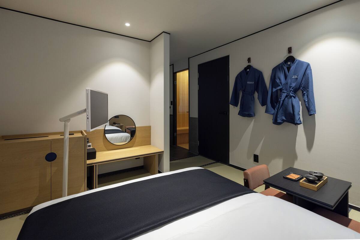 Quarto de hotel moderno com cama, mesa de cabeceira, espelho e dois roupões pendurados na parede.