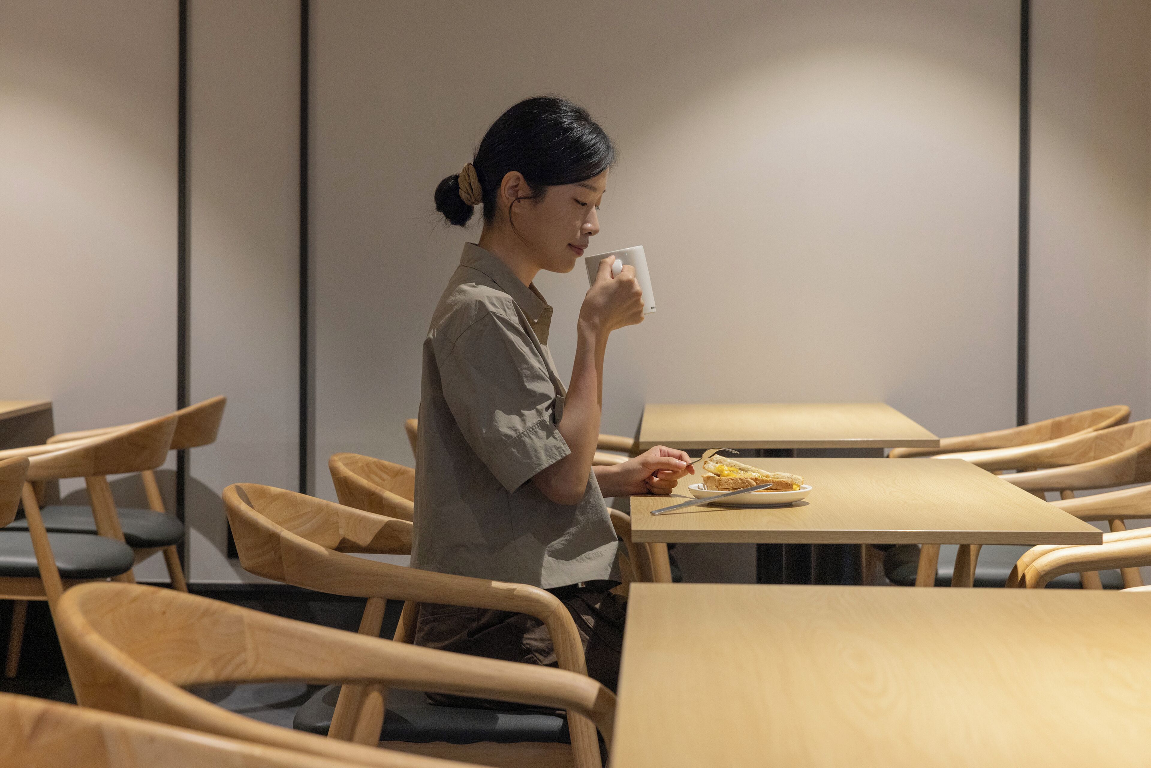 A person seated at a wooden table with a plate of food and a white mug, in a room with wooden chairs and tables.
