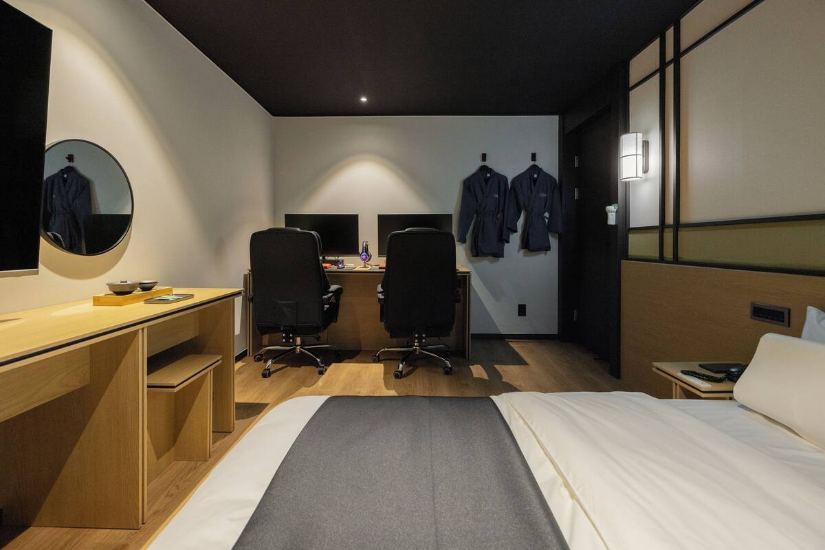 Quarto de hotel moderno com cama, escrivaninha, duas cadeiras, espelho e guarda-roupa.