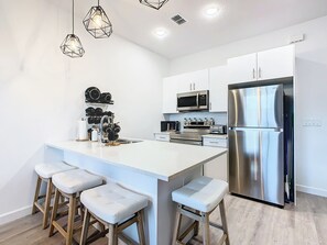 Una cocina moderna con armarios blancos, electrodomésticos de acero inoxidable, una isla central con taburetes altos, iluminación colgante y un microondas sobre la estufa.
