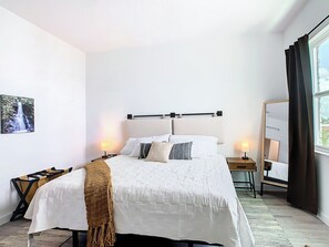 Una habitación de hotel con cama, mesitas de noche, un espejo, una ventana con cortinas y un cuadro colgado en la pared.