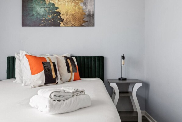 A neatly made bed with pillows, a folded towel, a green upholstered headboard, a bedside table with a lamp, and a painting on the wall.