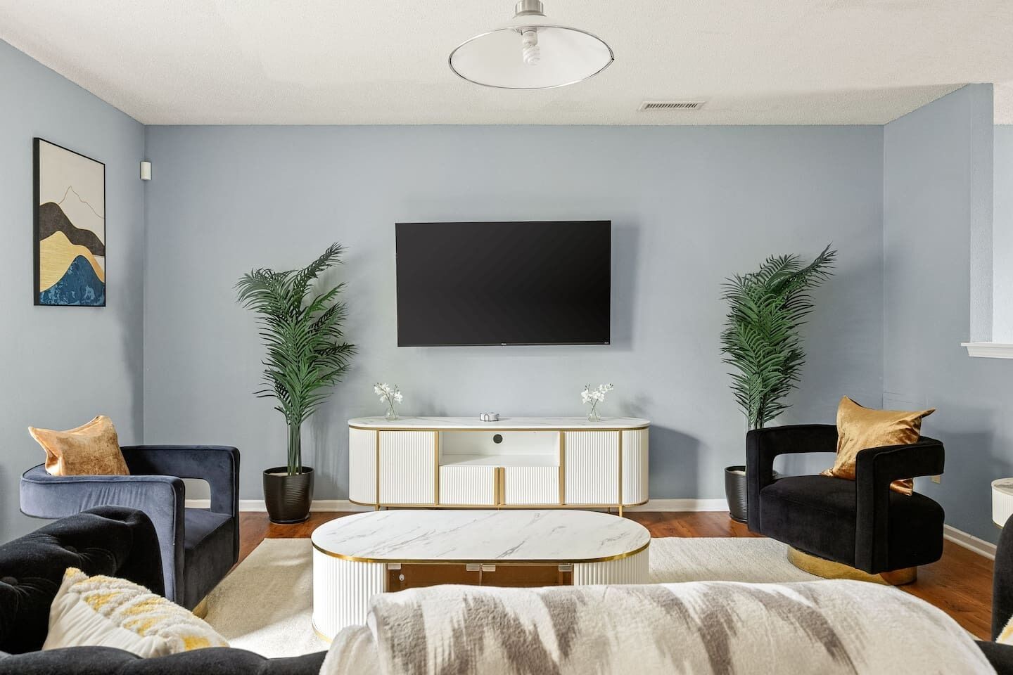 Un salon au design moderne, comprenant un grand téléviseur à écran plat, deux plantes en pot, une console blanche et deux fauteuils noirs agrémentés de coussins dorés.