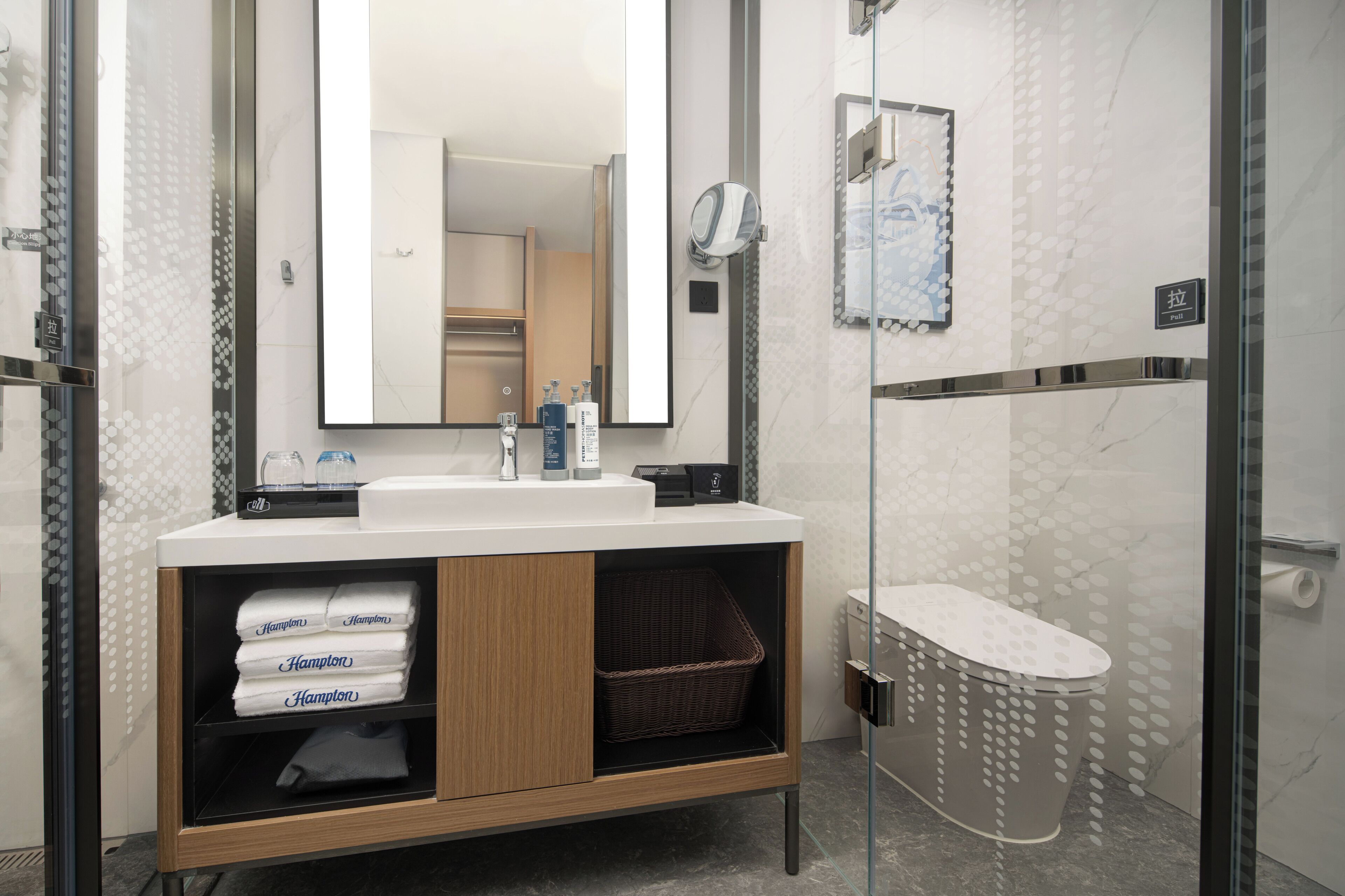 A modern bathroom with a large mirror, a sink, and a shower. There are towels and toiletries on the counter.
