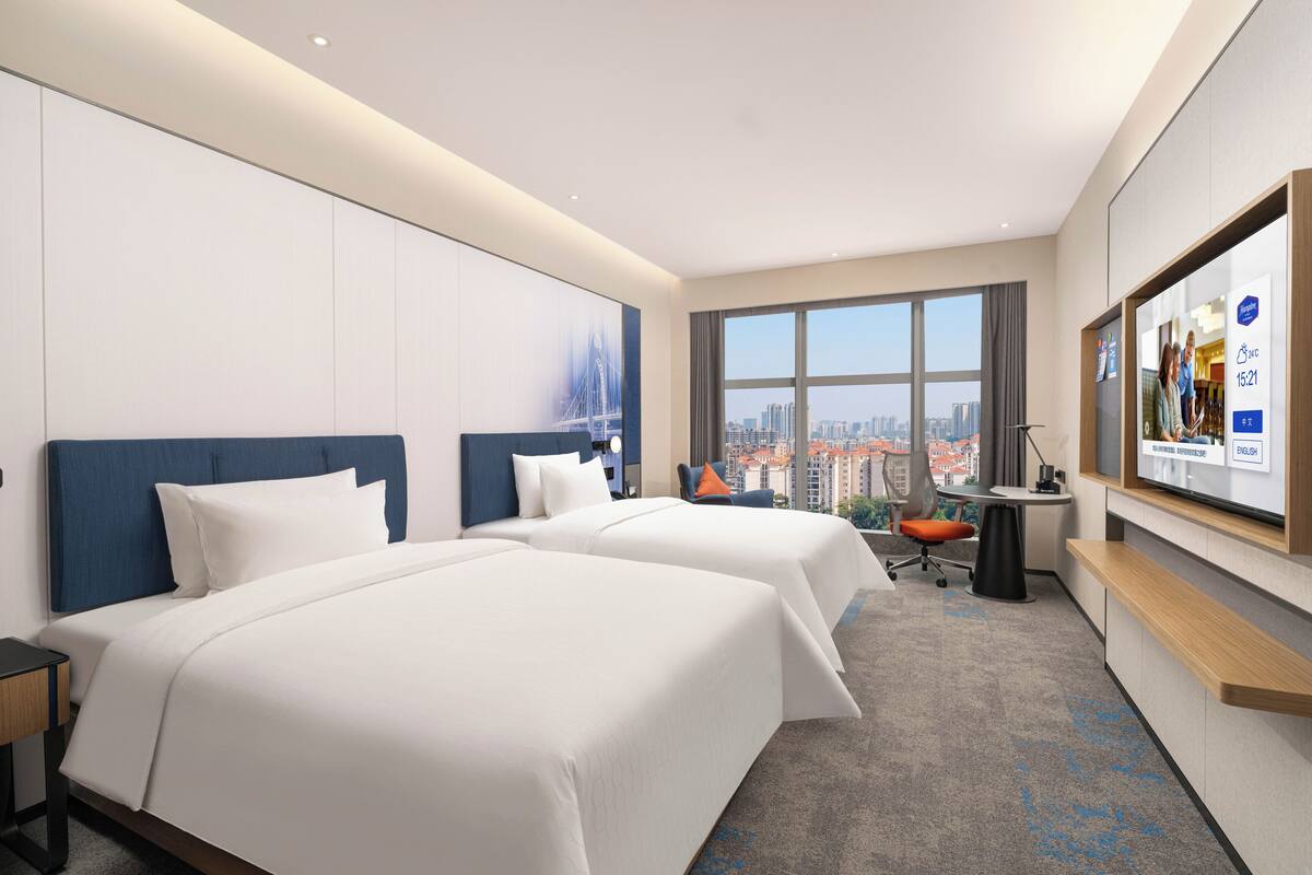 Habitación de hotel con dos camas, un escritorio, una silla, un ventanal amplio con vista a la ciudad y un televisor de pared.