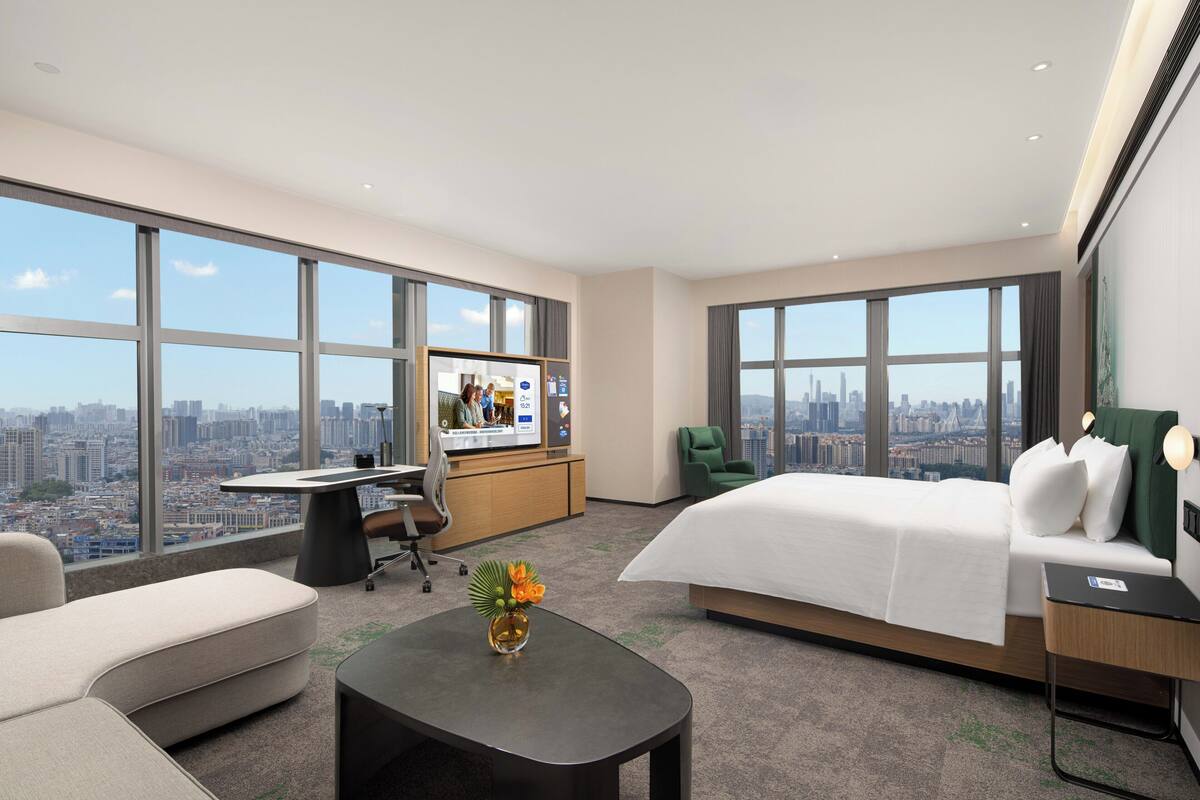 Habitación de hotel con cama, escritorio, silla, mesa de centro y ventanas amplias con vistas a la ciudad.