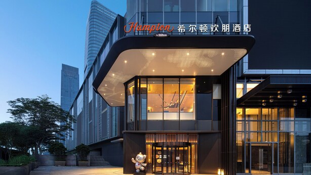 En moderne hotelindgang med en glasfacade, et synligt lobbyområde og et skilt, der angiver, at det er et Hampton hotel.