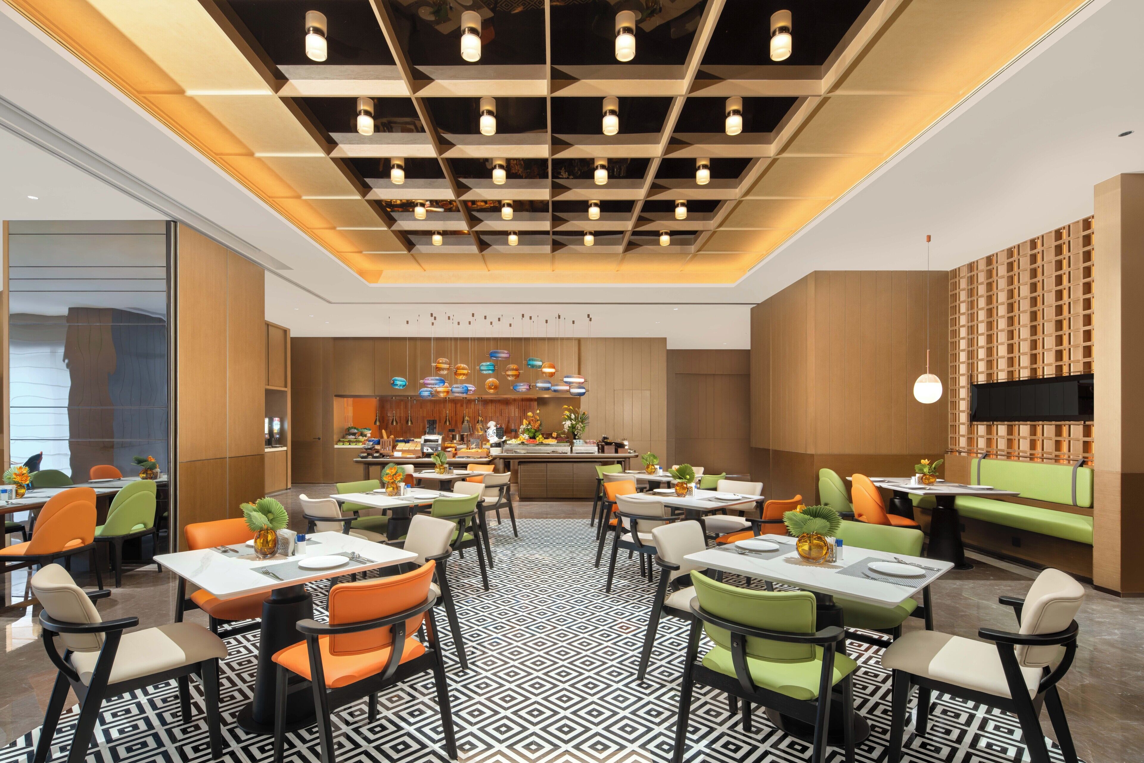 A modern restaurant with a geometric patterned floor, multiple dining tables with chairs, a central bar area, and a TV mounted on the wall.