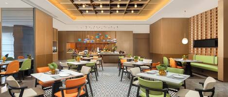 A modern restaurant with a geometric patterned floor, multiple dining tables with chairs, a central bar area, and a TV mounted on the wall.