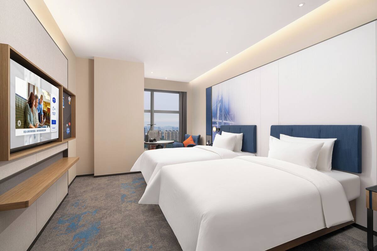Una habitación de hotel con dos camas, un televisor en la pared, una ventana con vistas a la ciudad y una pared de acento azul.