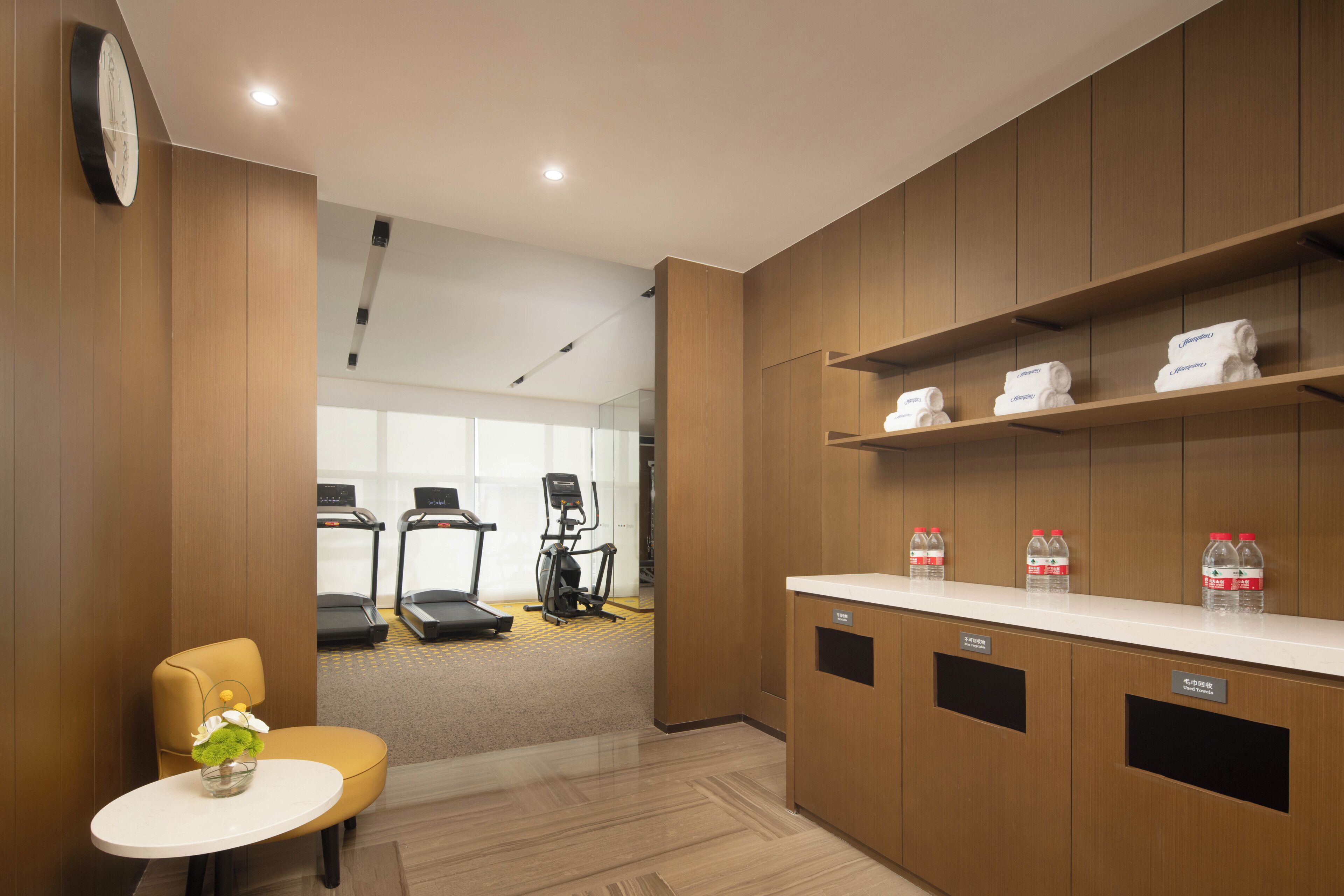 A hotel room with a wooden cabinet, a small table with a vase, a treadmill, and an elliptical machine in the background.