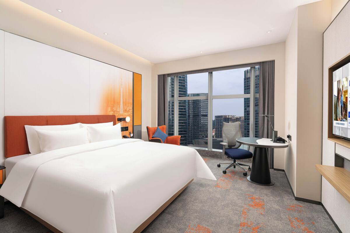 Habitación de hotel con una cama grande, un escritorio, una silla y un amplio ventanal con vistas a la ciudad.