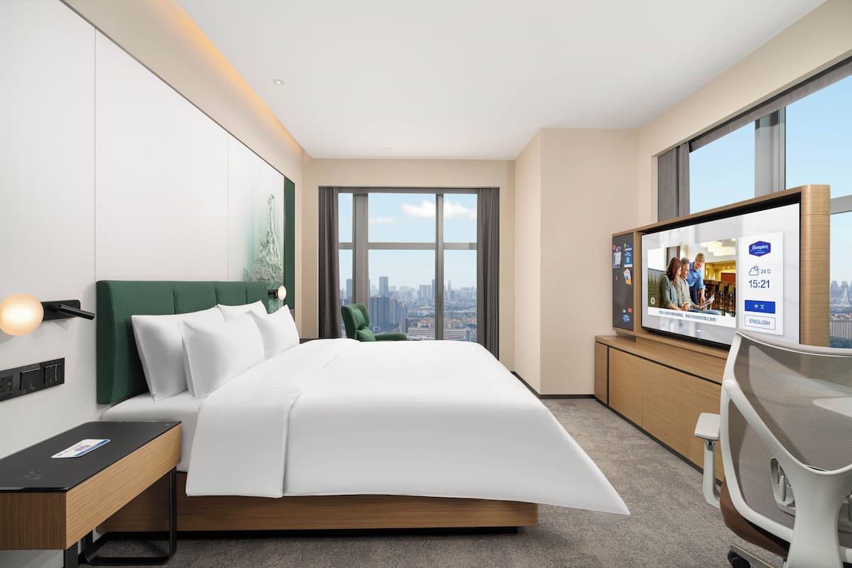 Habitación de hotel moderna con cama, escritorio, silla, televisor y un amplio ventanal con vistas a la ciudad.
