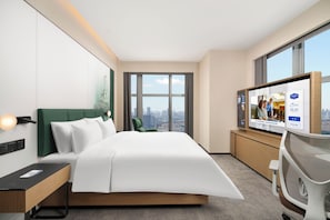 Une chambre d’hôtel moderne équipée d’un lit, d’un bureau, d’une chaise, d’une télévision et d’une grande fenêtre donnant sur la ville.