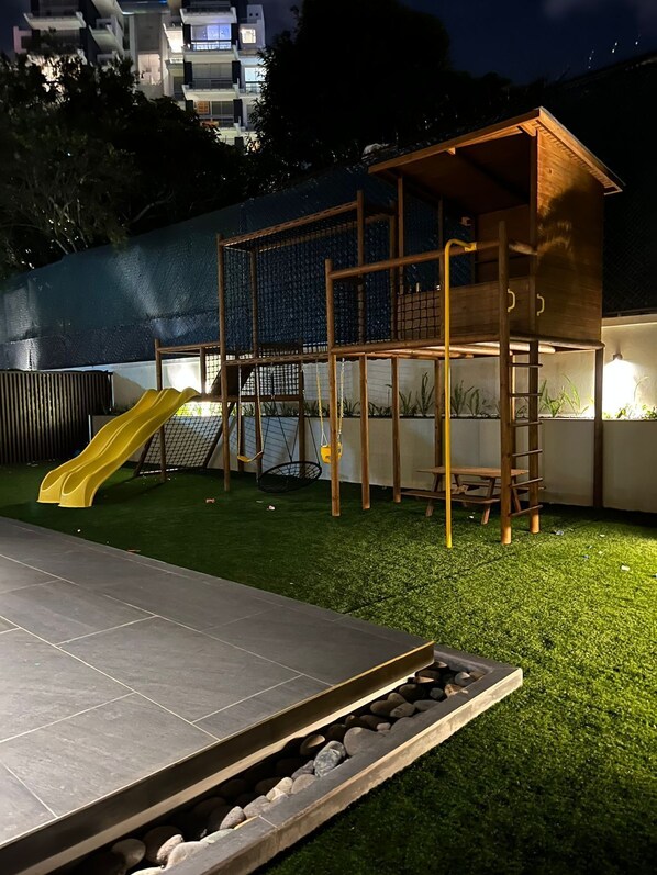 A playground with a slide and climbing structures, surrounded by artificial grass and a stone border. - Luxurious Apt. Z.10 GT City | 2Bedroom | AC | Wifi | Parking | Swimming Pool Jacuzzi (Ciudad de Guatemala)