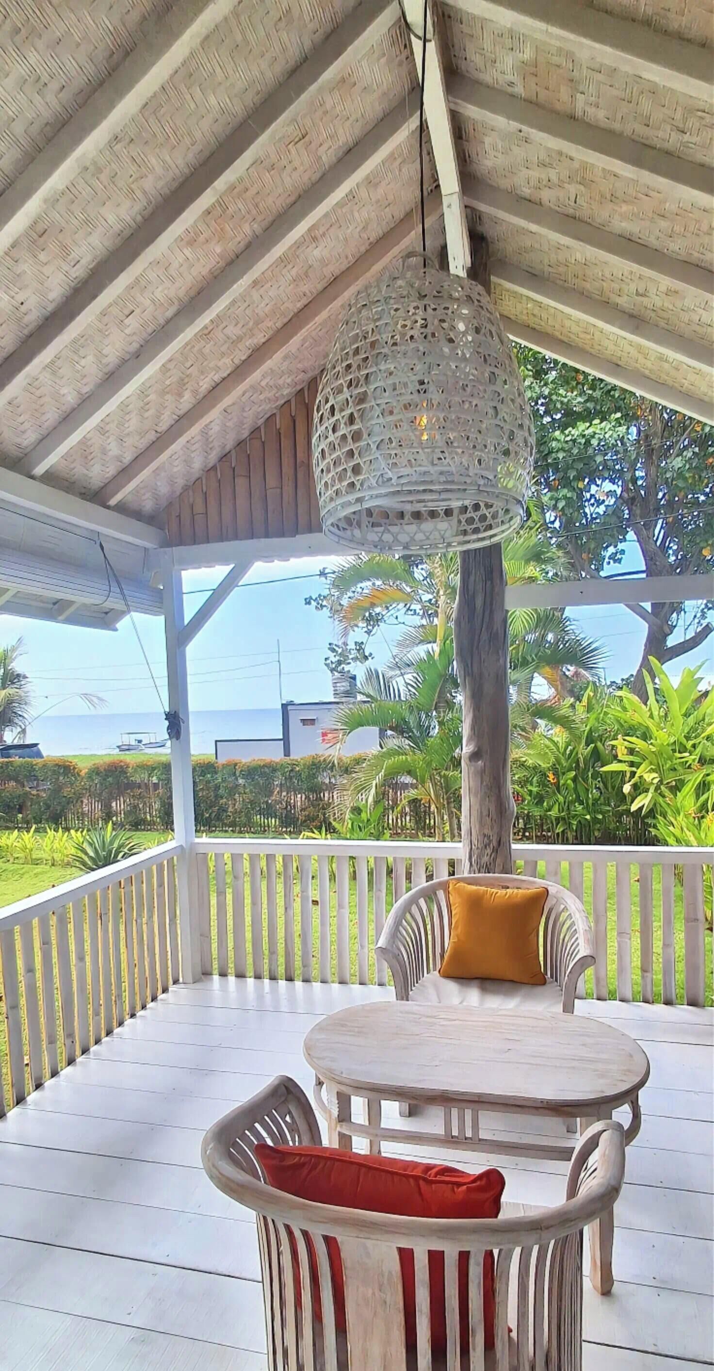 A covered outdoor seating area with a woven ceiling light fixture, white wicker furniture, and a view of the ocean.