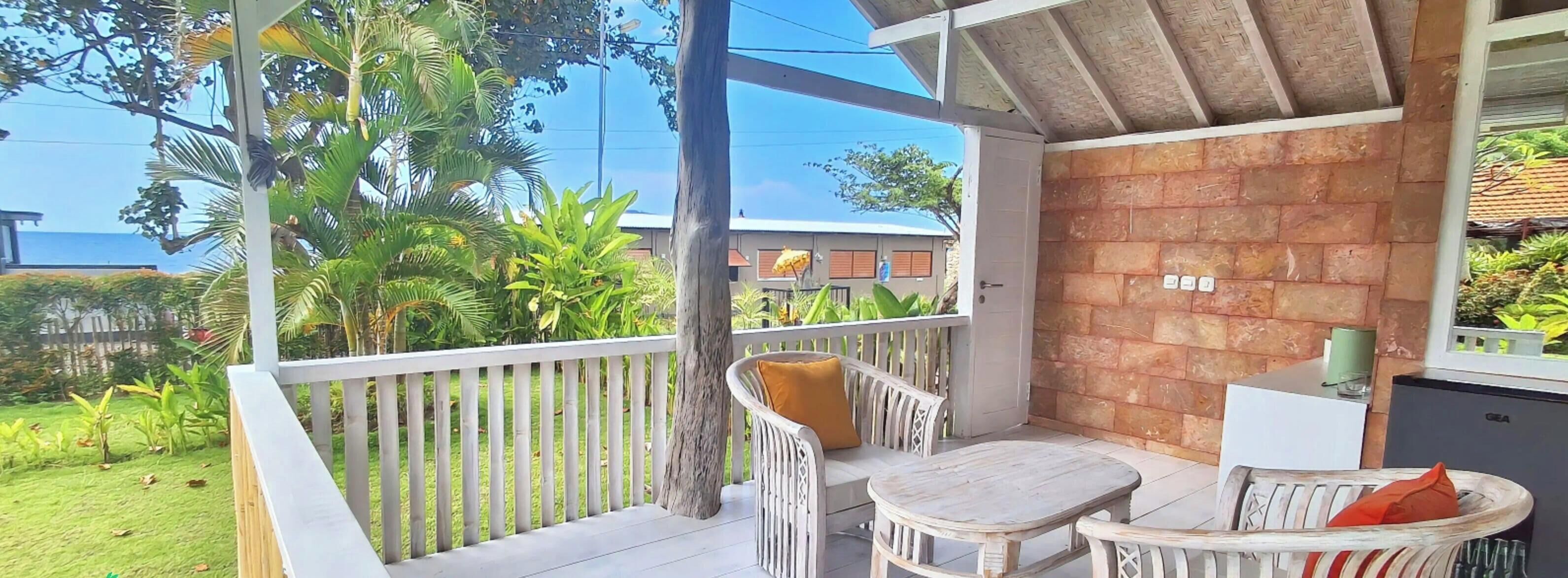 A balcony with a white railing, a white wicker sofa with a yellow cushion, a wooden table, and a wicker chair with an orange cushion.