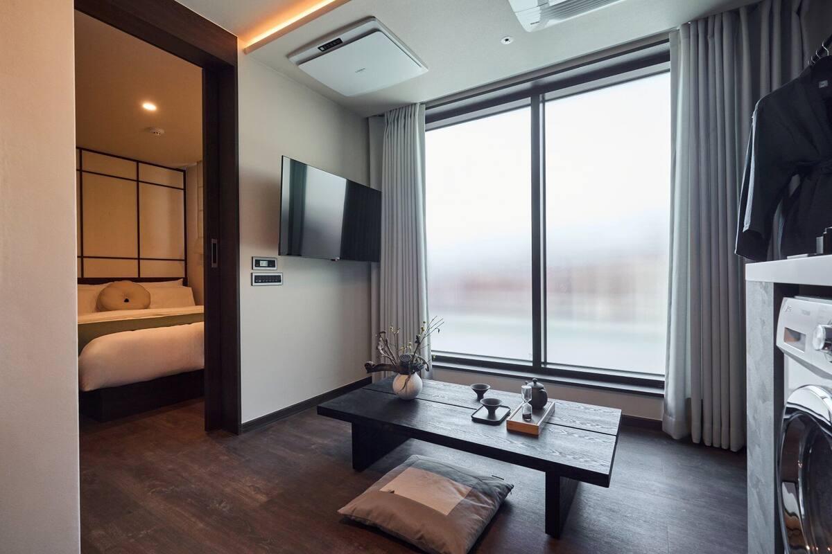 Une chambre d’hôtel moderne équipée d’un lit, d’un téléviseur à écran plat, d’une grande fenêtre avec des rideaux, d’une table basse en bois et d’une table de chevet agrémentée d’un vase.